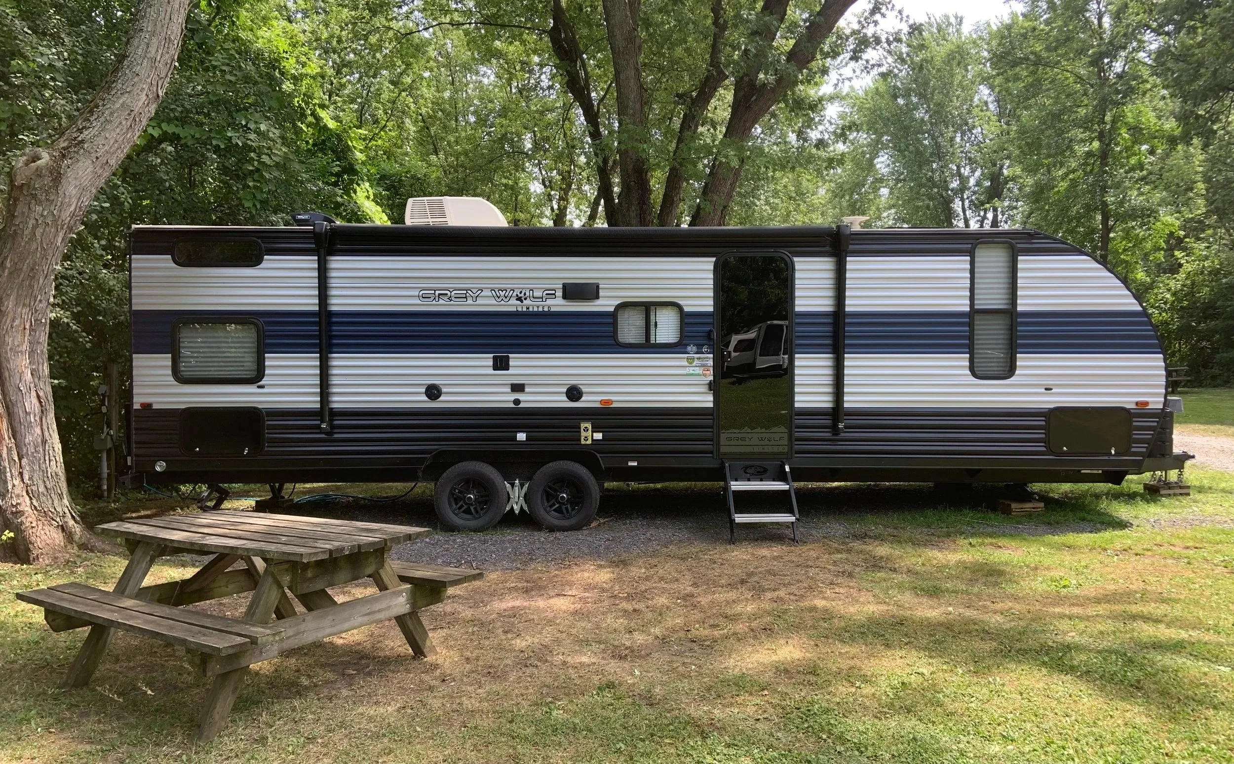 A grey and black Grey Wolf travel trailer parked on a patch of grass and gravel, surrounded by trees. There's a small wooden picnic table in front of it and a set of steps leading to the door.