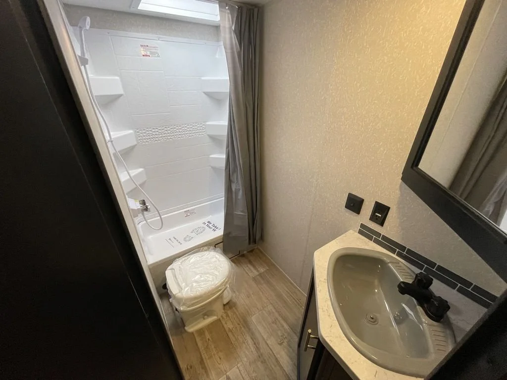 Small bathroom with a shower stall, toilet, and sink with a mirror above. The shower has a curtain, and the tiles are white with textured shelves. The sink is set in a dark cabinet with a countertop.