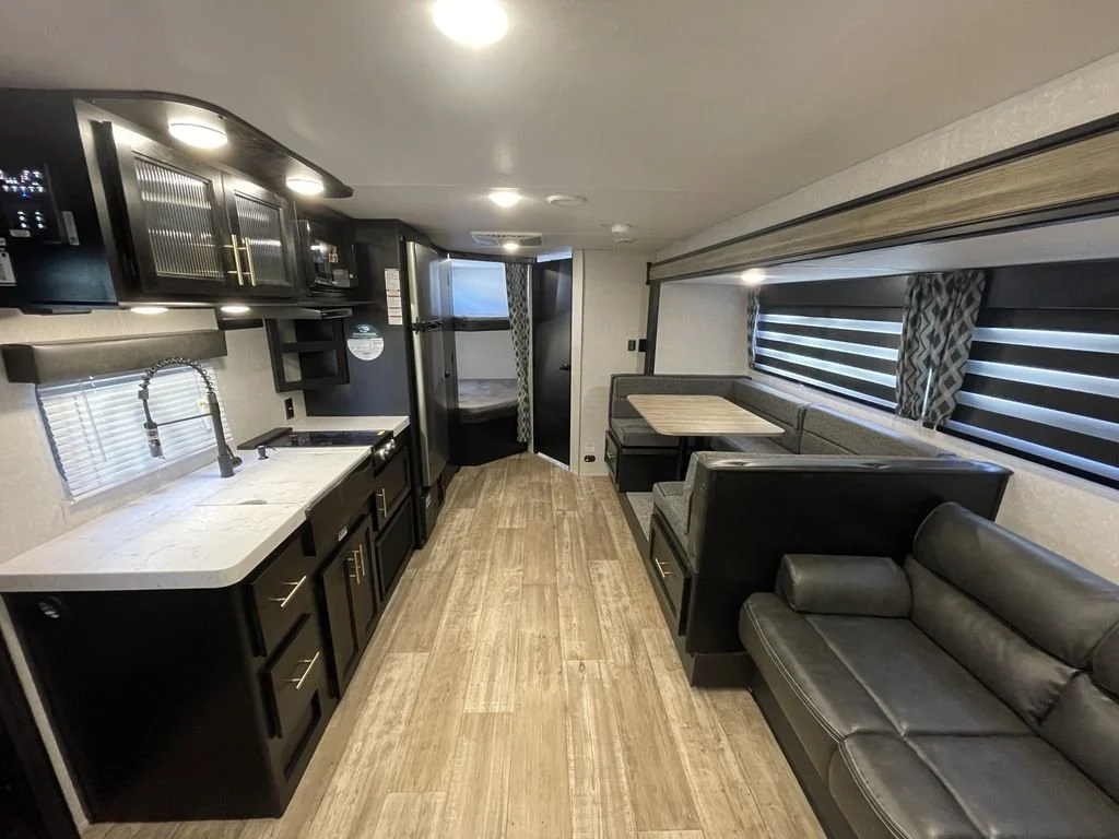 Interior of a modern RV with a kitchen area, dining nook, and sofa, featuring black cabinets, wood flooring, and striped window blinds.