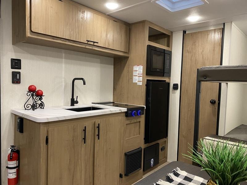 Compact kitchen area with wood cabinets, black microwave, mini fridge, electric stove, black faucet, black vent, and decorative candles on the countertop, in a small living space.
