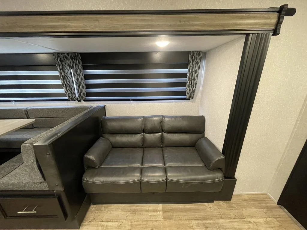A black leather sofa with armrest pillows, placed against a white textured wall beneath two windows with horizontal black and gray striped blinds and patterned curtains.