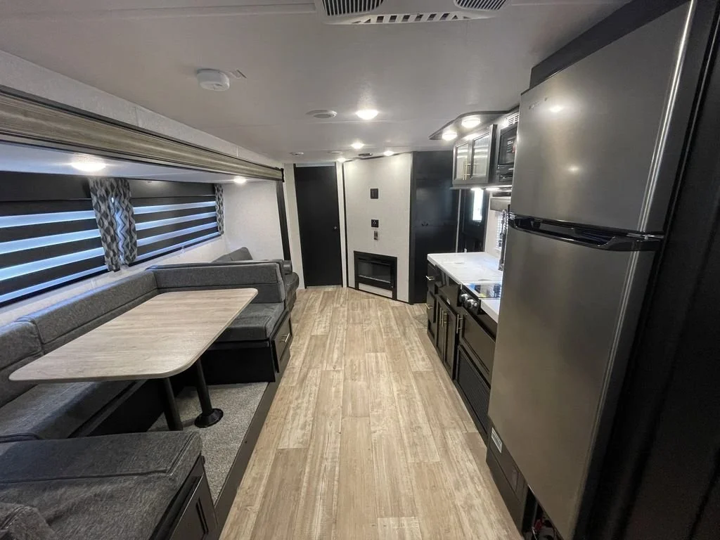Interior of a modern RV with a dining area on the left, kitchen appliances on the right, and windows with striped blinds.