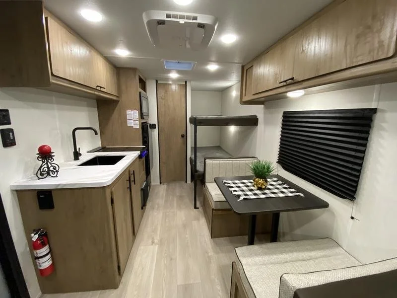 Interior of a compact kitchen and dining area in an RV with wood cabinets, black window shades, a small dining table with a checkered tablecloth, and a kitchenette with a sink and appliances.
