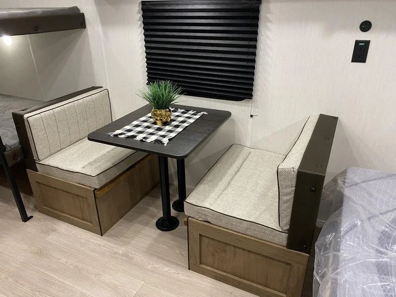 A small dining nook with a black table, two beige upholstered bench seats, a black and white checkered table runner, and a green plant in a gold pot on the table. There is a window with black blinds behind the table.