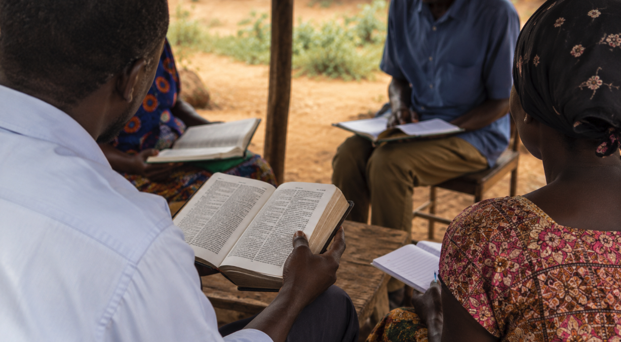 Group of people sitting outdoors, reading and discussing from books and notebooks during a gathering.