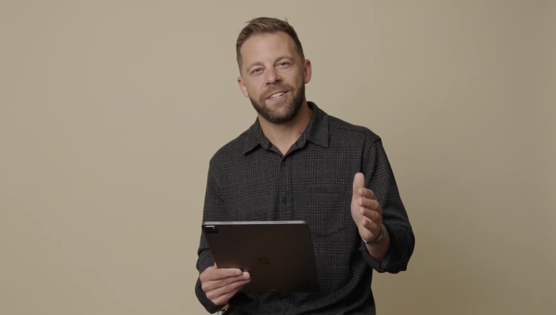 A man with light brown hair and a beard, wearing a dark checkered shirt, is gesturing with his right hand while holding a tablet in his left hand, standing against a plain beige background.