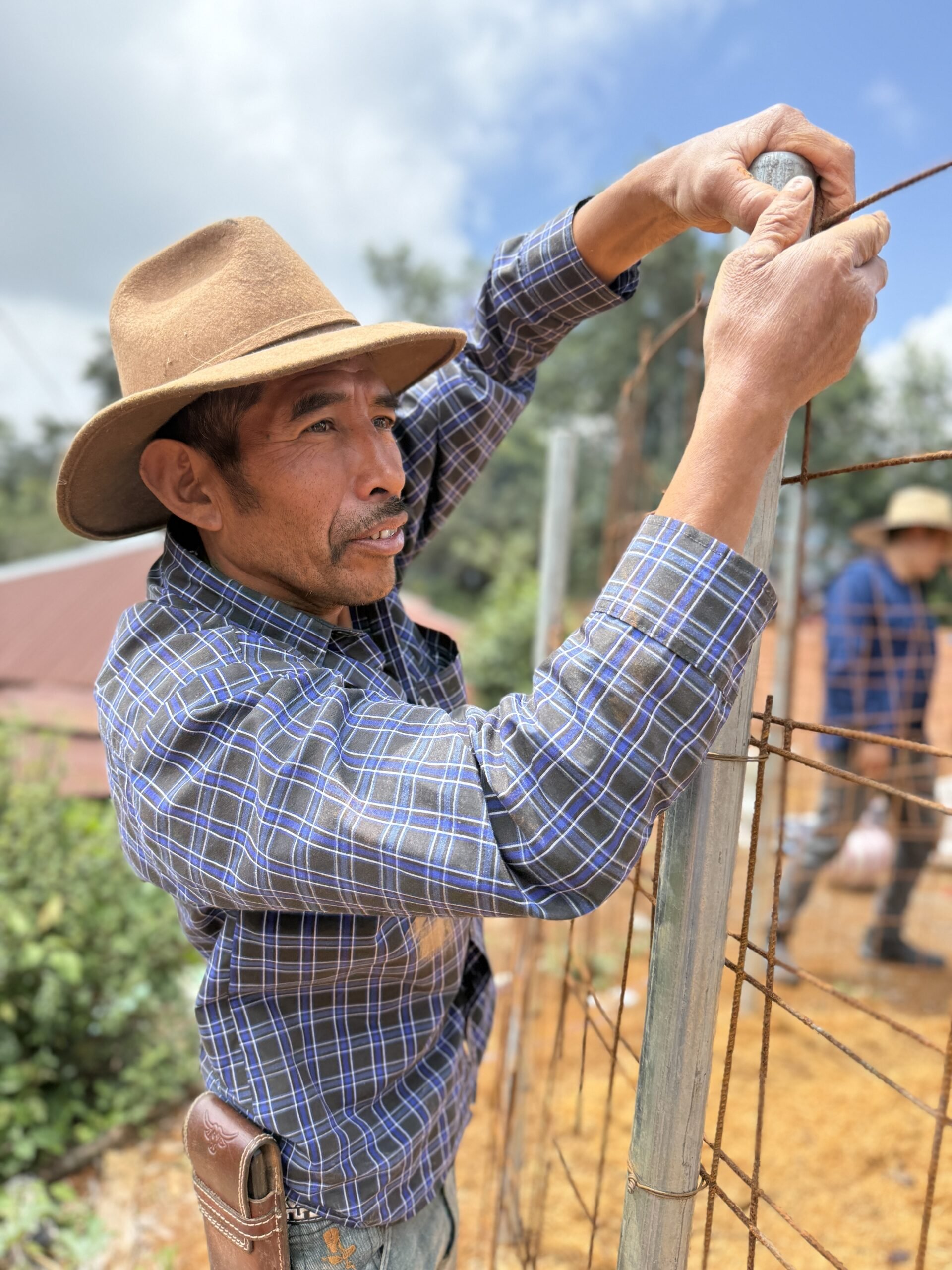 A man wearing a wide-brimmed hat and a plaid shirt is working outdoors on a wire fence, using a tool to secure the wire to a post, with another person blurred in the background.
