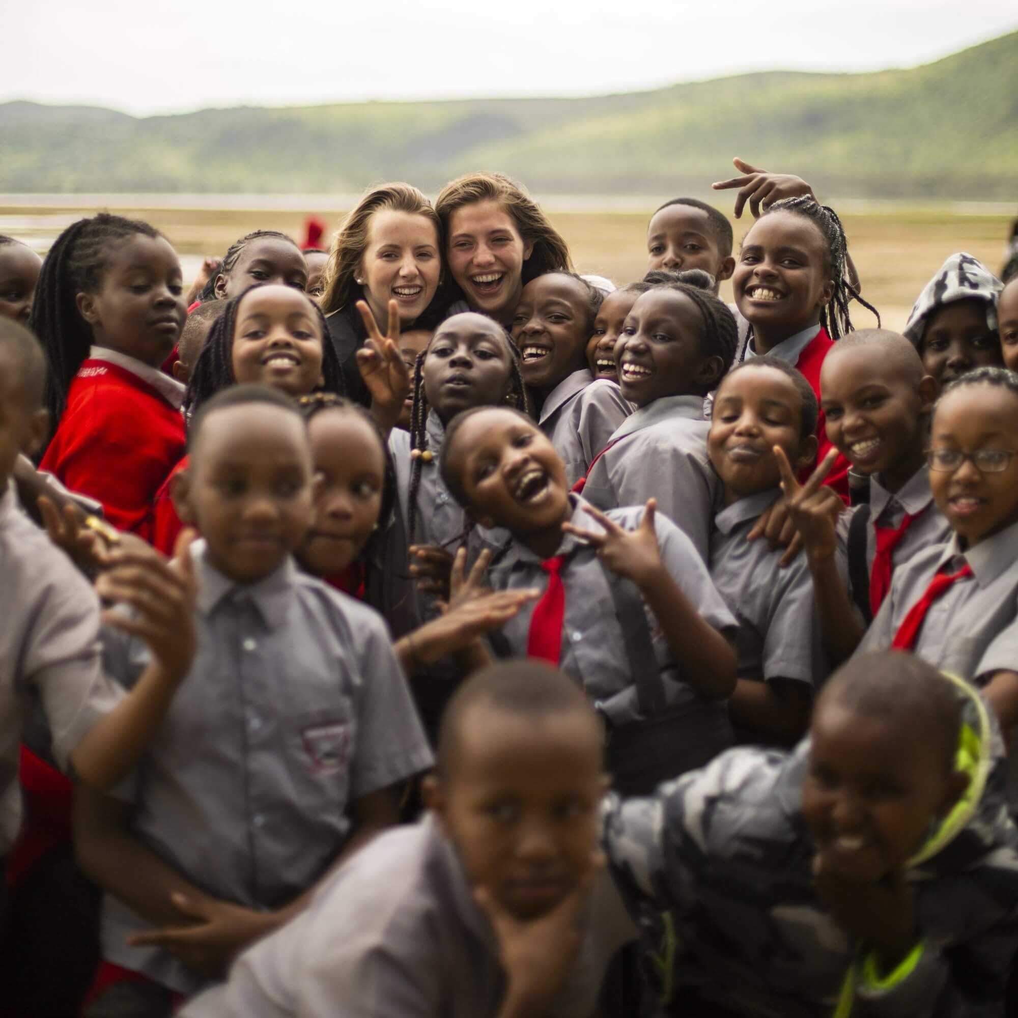 A group of smiling children and two women taking a group photo outdoors against a hilly landscape in the background.