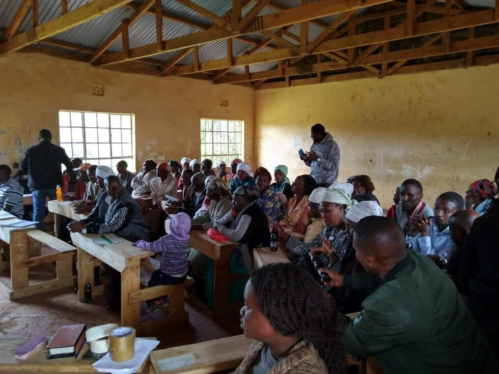 A large group of people gathered in a rustic indoor setting with yellow walls and a metal roof, attending a meeting or workshop, with some individuals taking notes or using electronic devices.