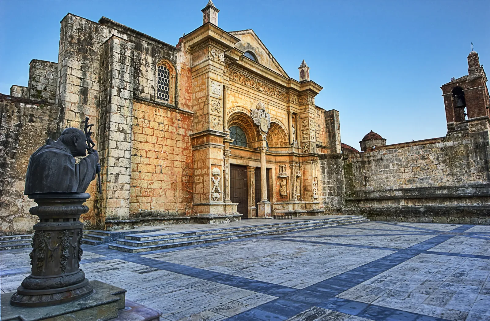 A historical building with intricate architectural details, stone walls, and a wooden door, surrounded by a stone courtyard. There is a bronze bust of a man on a pedestal in the foreground and a clock tower in the background.