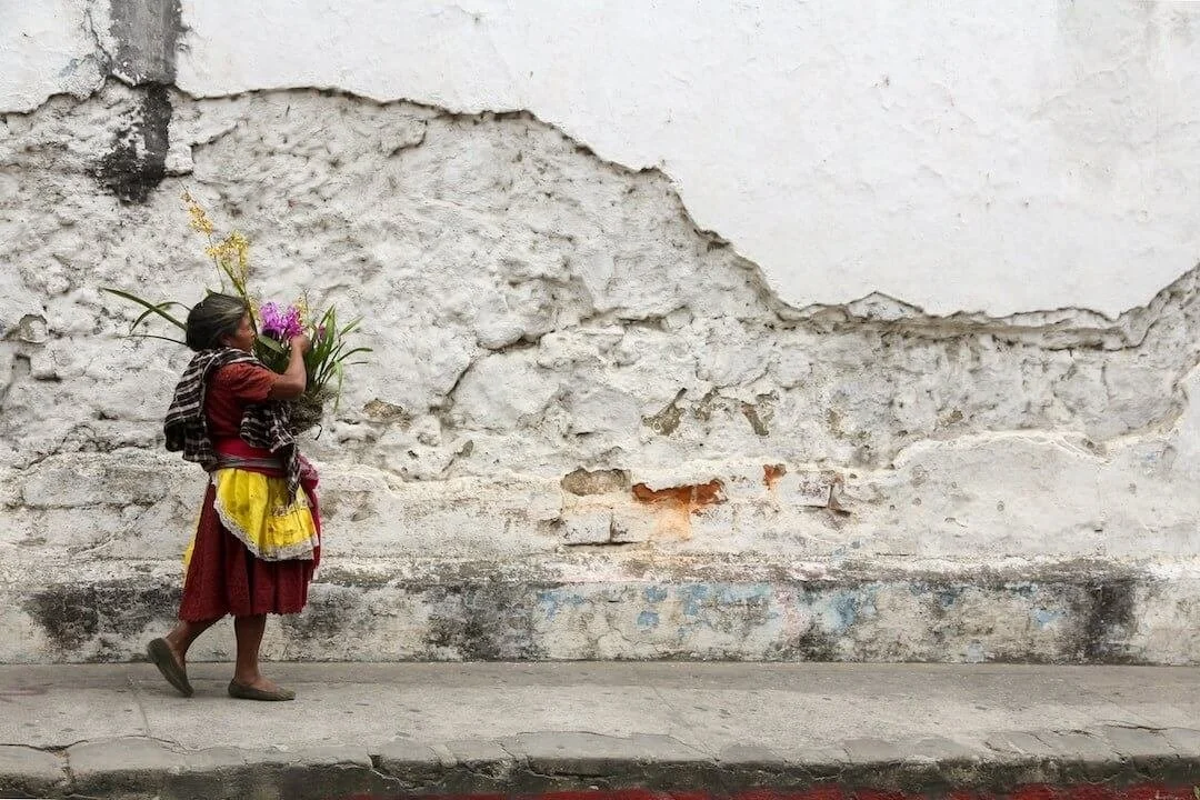 A woman wearing traditional clothing walking along a worn brick sidewalk against a textured, weathered white wall, carrying a bouquet of flowers.