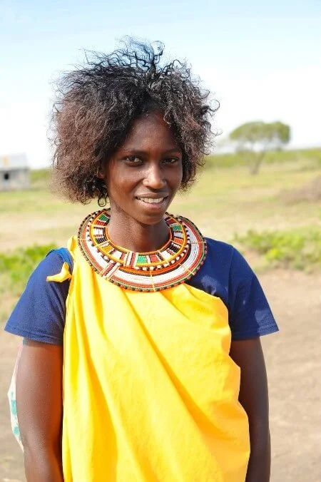 Woman in traditional attire with colorful beaded necklace, standing outdoors in a rural area with green fields and a tree in the background.