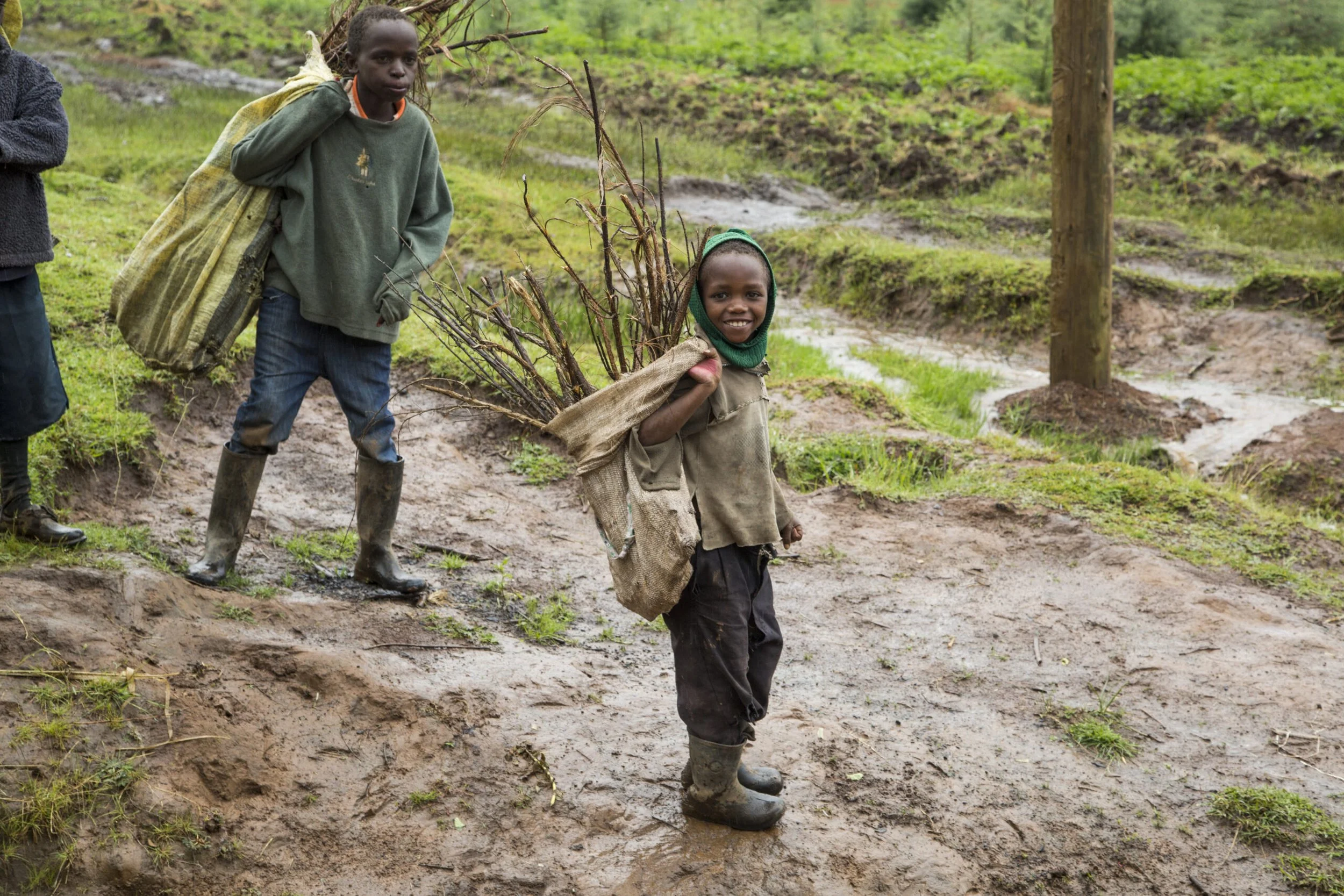 Two children walking through a muddy trail in a green, wooded area. One child is carrying a bundle of sticks on their shoulder, smiling and wearing a hoodie and jacket. The other child, wearing boots and a coat, looks serious. In the background, the landscape is lush with grass and trees.