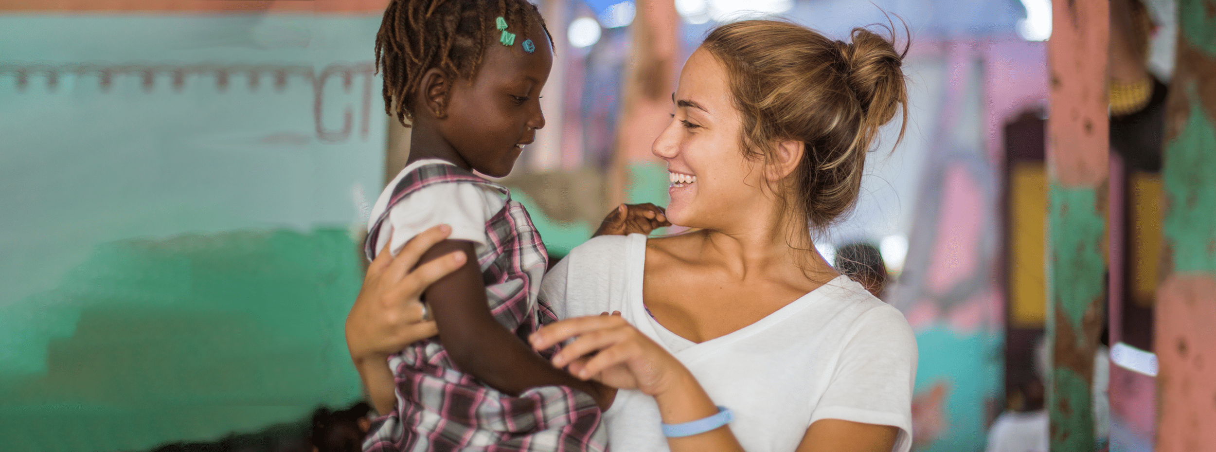 A woman with light brown hair smiling and holding a young girl with dark skin, dreadlocks, and wearing a plaid shirt in a colorful indoor setting.