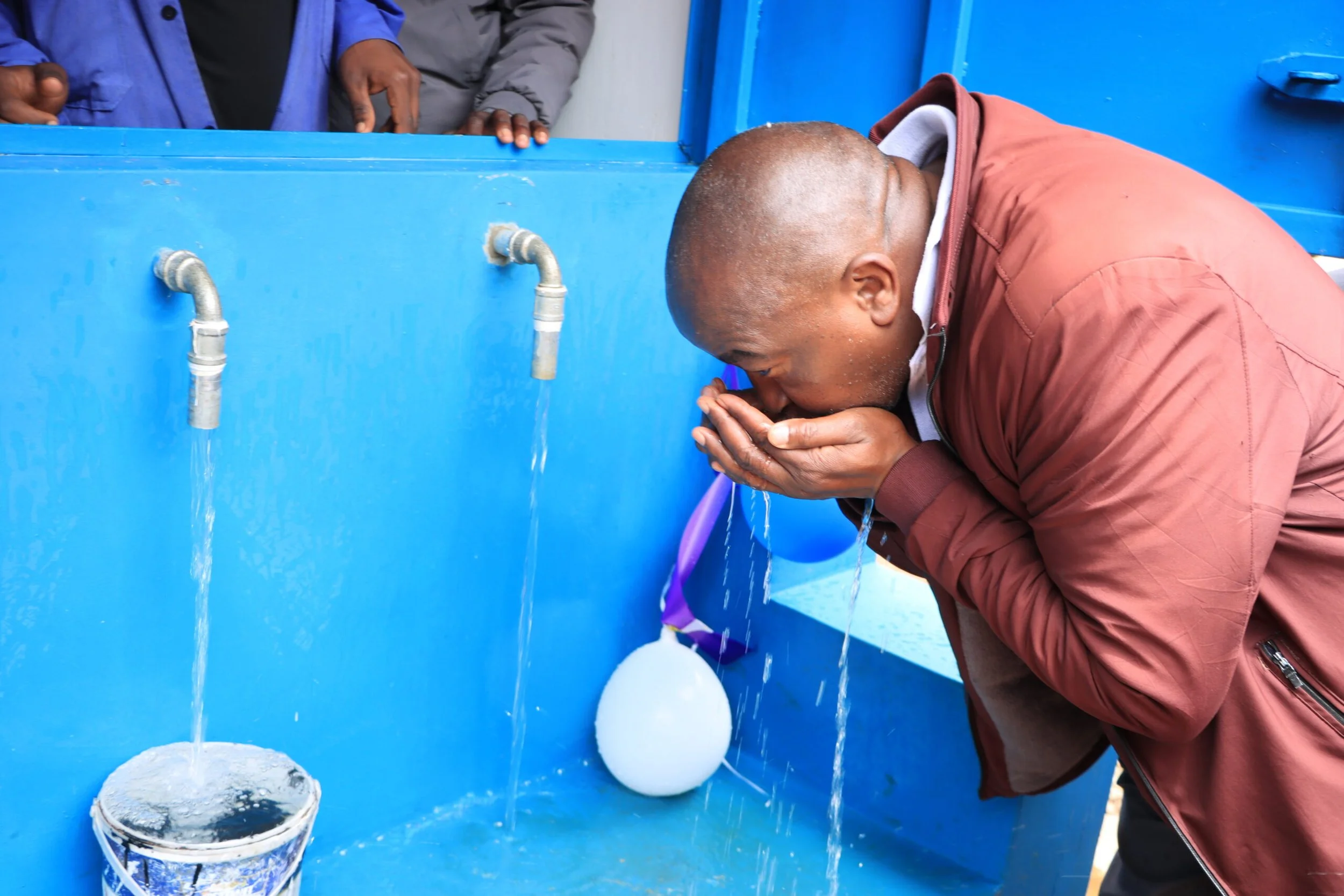 Man with a shaved head leaning over a blue sink, washing his face with water from the tap.