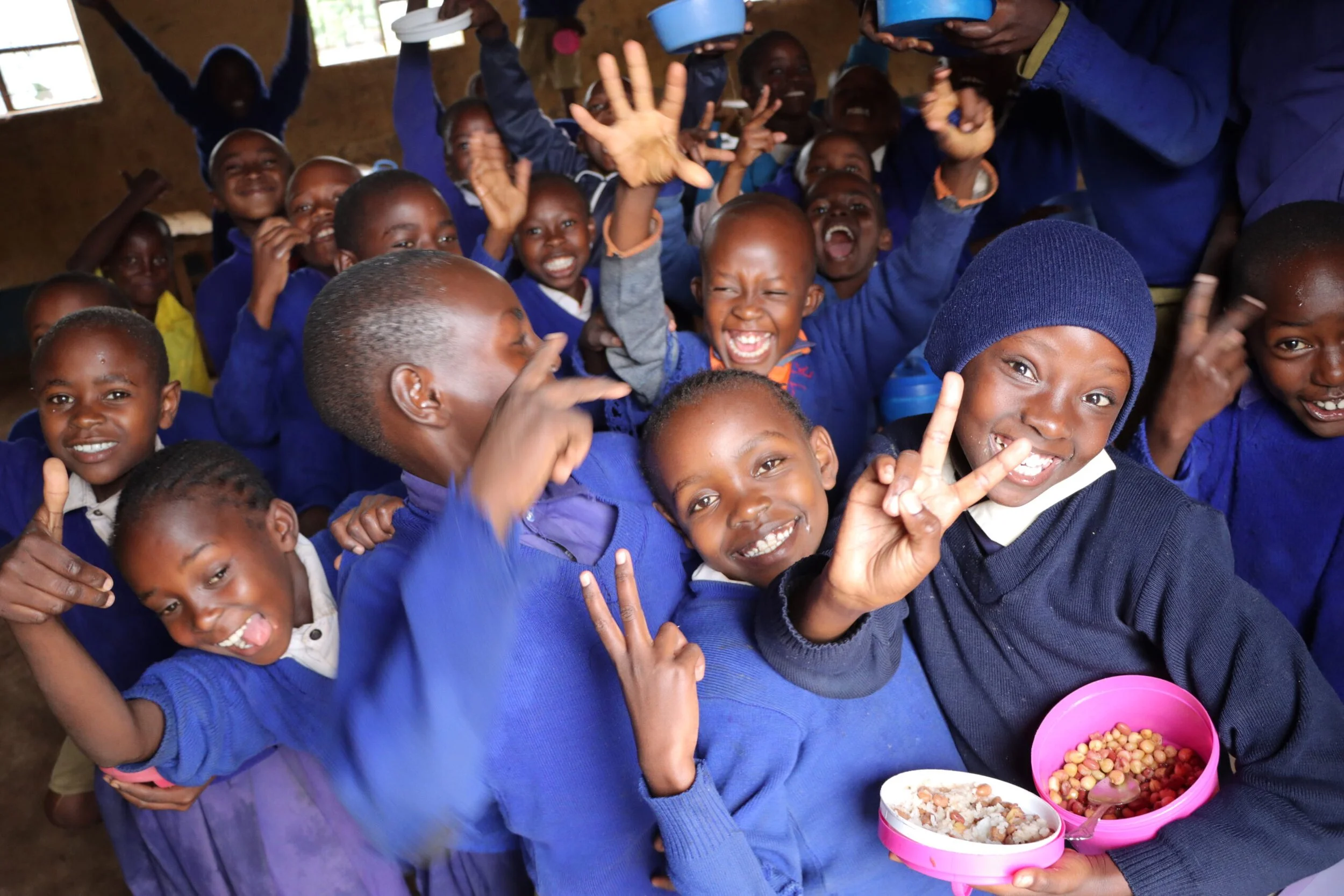 A group of children in blue clothing smiling and enjoying a meal together inside a simple building with small windows, some children making peace signs and others laughing.
