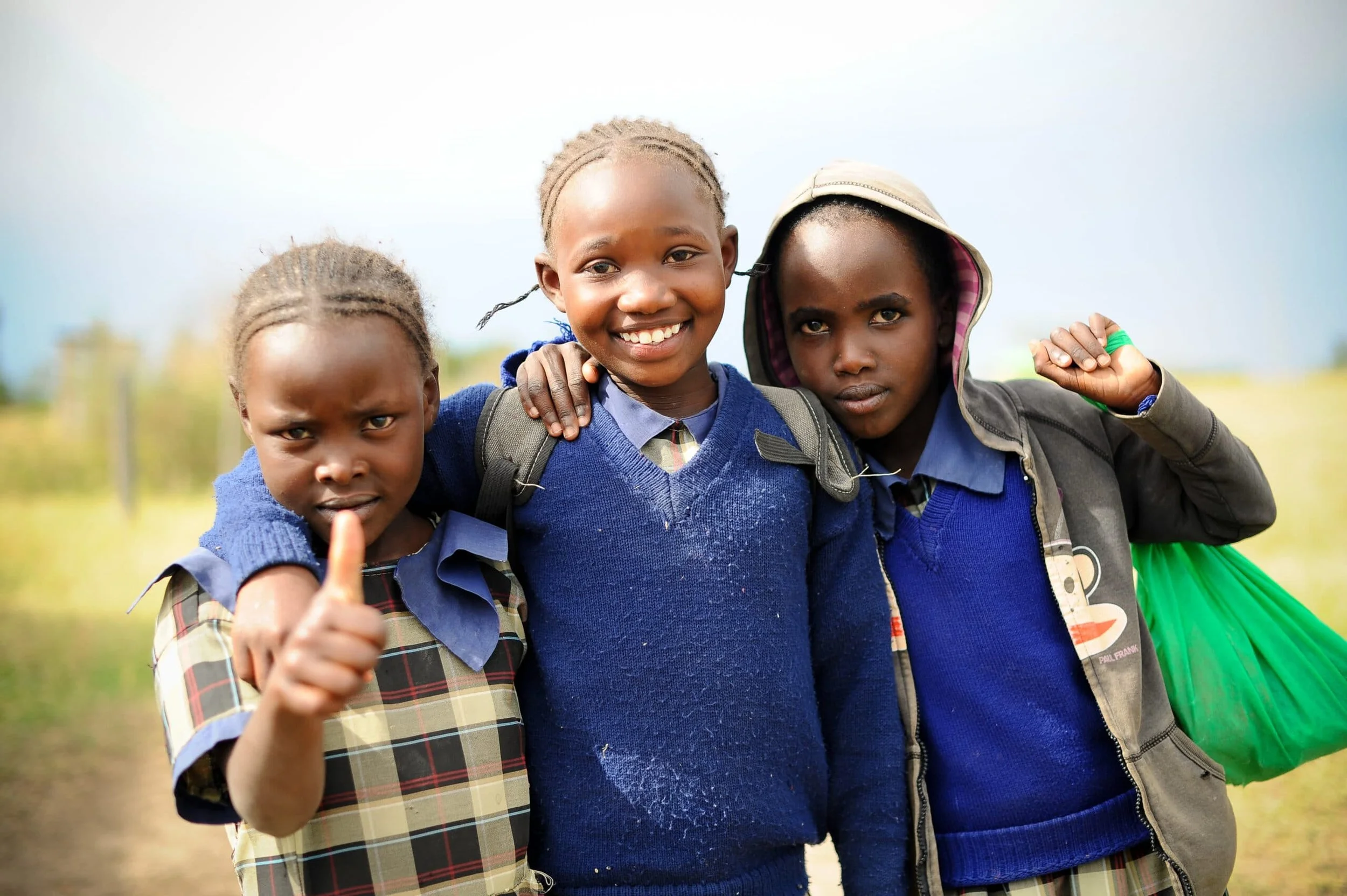 Three young girls outdoors, smiling and wearing school uniforms, with one giving a thumbs-up gesture.
