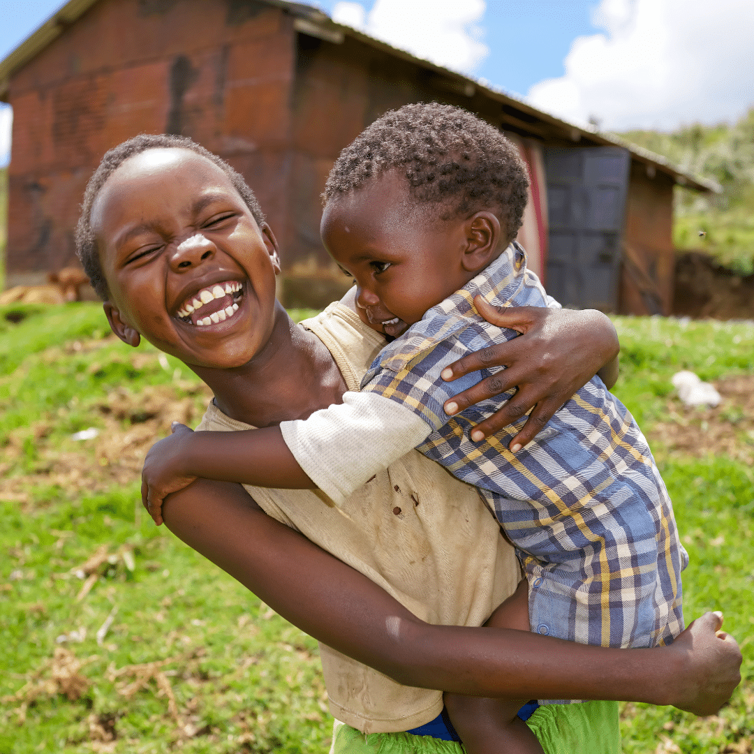 Two boys hugging and smiling joyfully outdoors in front of a rustic building with a grassy area.