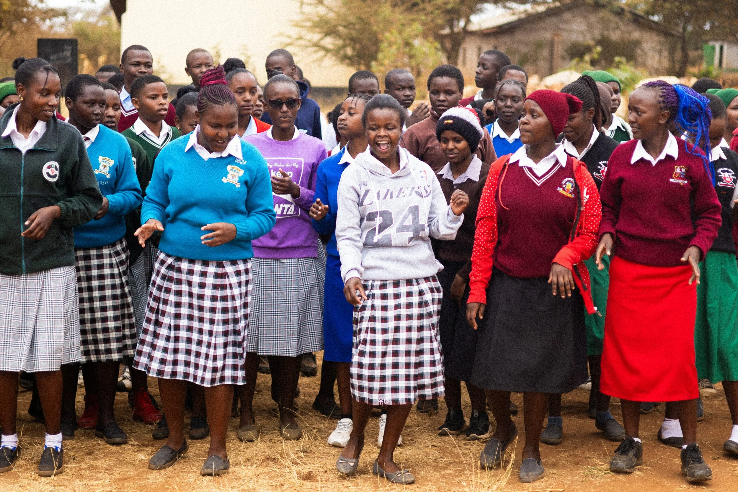 A group of students and a teacher are walking outdoors, some smiling and talking, in a school uniform setting, with trees and buildings in the background.