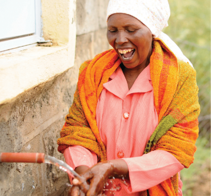 A woman smiling and washing her hands with water outside a brick building, wearing a colorful shawl and pink blouse.