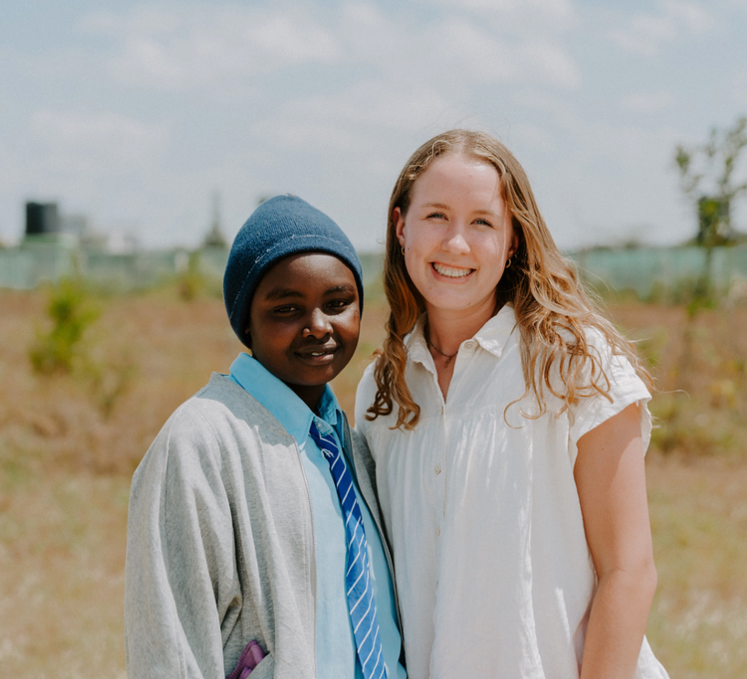 Two women standing outdoors, smiling at the camera. One is a young woman with long blond hair in a white shirt, and the other is a girl with dark skin, wearing a beanie, a light blue shirt, a blue tie, and a gray jacket.