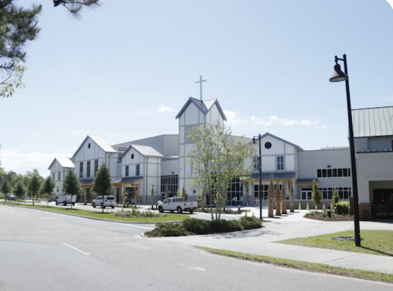 A modern church building with a steeple topped by a cross, surrounded by parking lot, parked cars, trees, lampposts, and a sidewalk on a clear, sunny day.