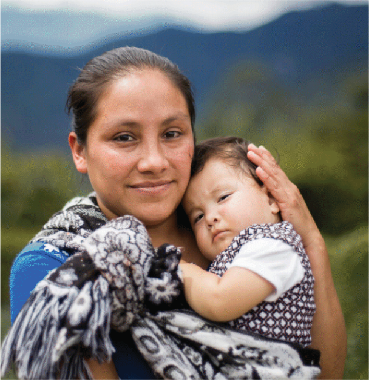 A woman holding a young child, outdoors with mountains and greenery in the background.