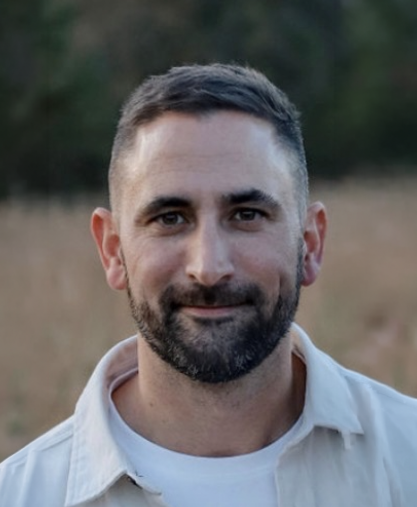 A man with short dark hair and a beard smiling outdoors in a field with blurred trees in the background.