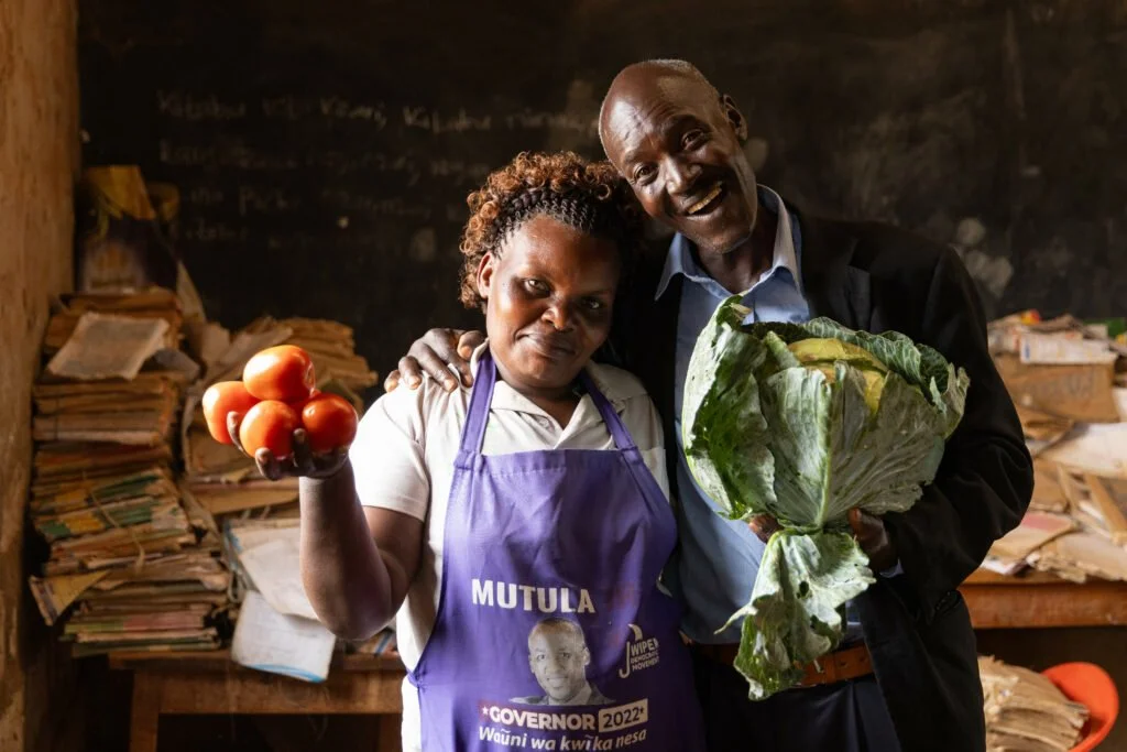 A woman wearing a purple apron and a man in a black jacket standing inside a room with stacks of papers or books behind them, holding tomatoes and a large leafy vegetable.