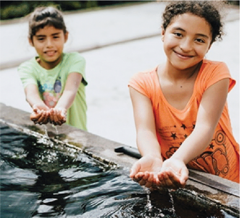 Two young girls filling their hands with water from a fish pond or water feature outdoors, smiling at the camera.
