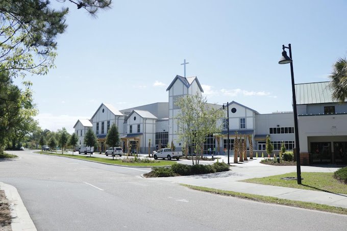 Exterior of a modern church and shopping center with parked cars, sidewalk, trees, and street lamps.