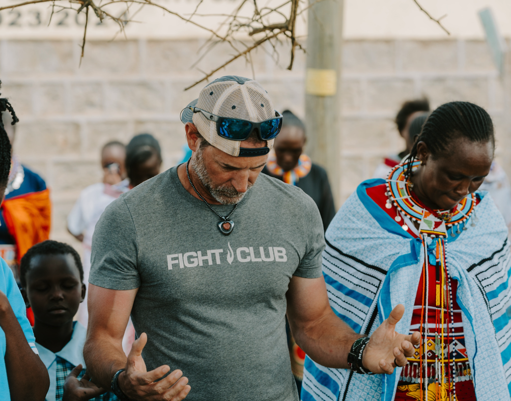 A man wearing a gray "Fight Club" t-shirt, a cap with sunglasses on top, and a necklace, praying or meditating with eyes closed, in a group of people in traditional Maasai attire outdoors.