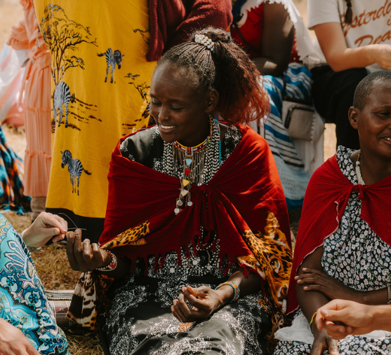 A woman with braided hair seated on the ground, wearing a red shawl and traditional jewelry, smiling and looking at her hands during an outdoor gathering with other people.