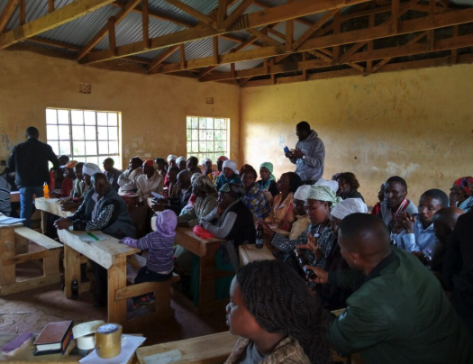 A group of people seated on wooden benches in a simple, rustic classroom with yellow walls and a metal roof, many taking notes or using their phones.