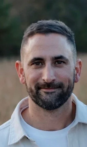 A man with short dark hair and a beard, smiling outdoors with a blurred background of trees.