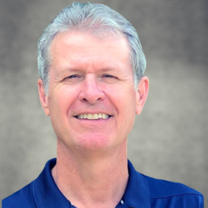A smiling man with gray hair and a blue collared shirt, standing outdoors against a neutral background.