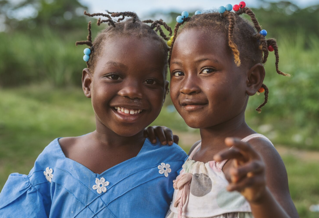 Two young girls with braided hair decorated with beads, smiling outdoors, one pointing towards the camera.