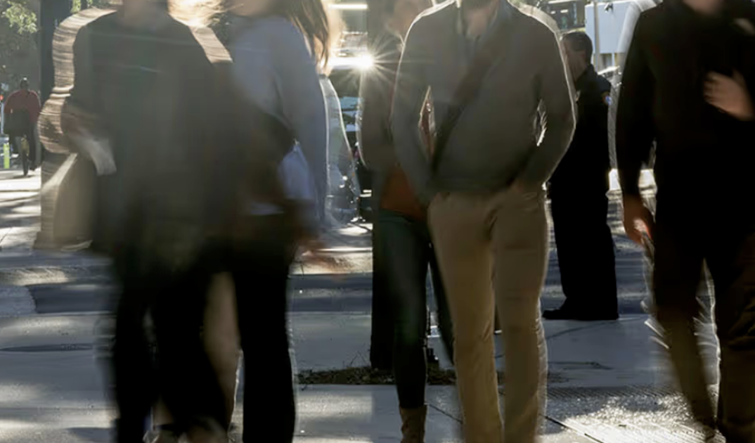 Group of people walking on a busy city sidewalk with sunlight glare in the background.