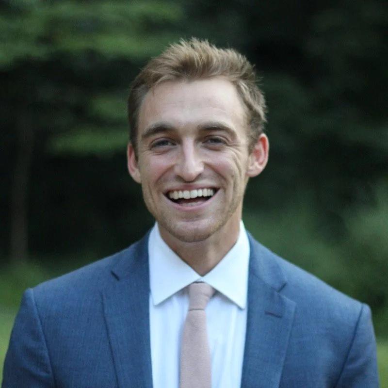 A smiling man in a blue suit, white shirt, and beige tie outdoors with trees in the background.