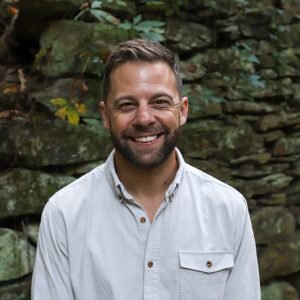 A smiling man with short hair and a beard, wearing a light-colored button-up shirt, standing outdoors against a rocky background.