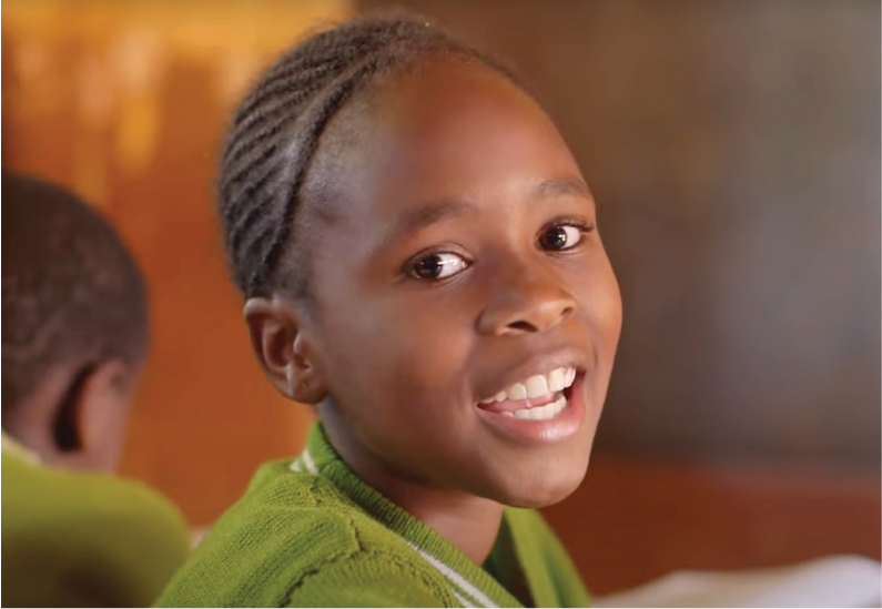 A young girl with braided hair smiling in a classroom setting, wearing a green sweater.