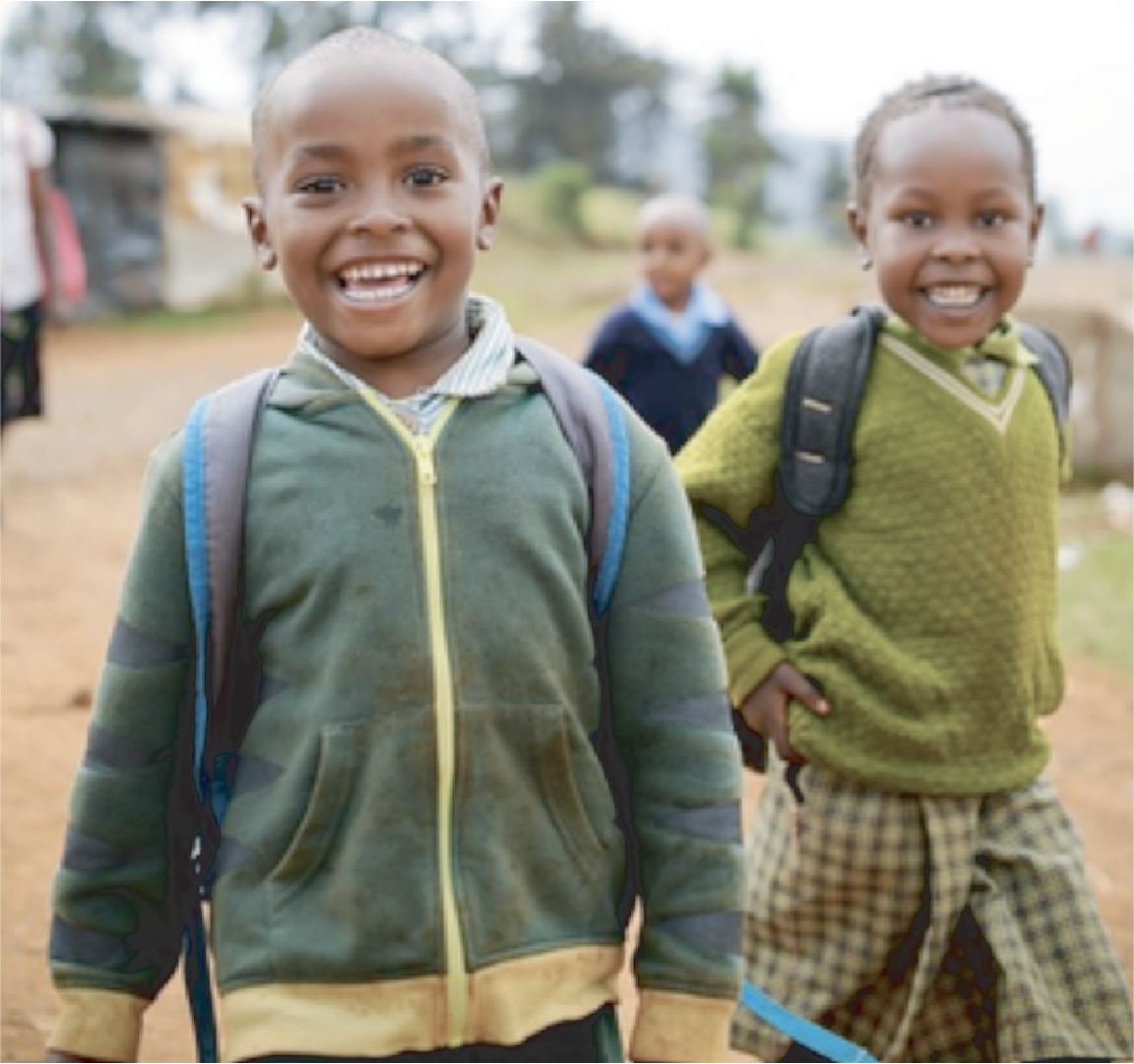 Two smiling children with backpacks standing outdoors in a rural setting, with another child in the background.