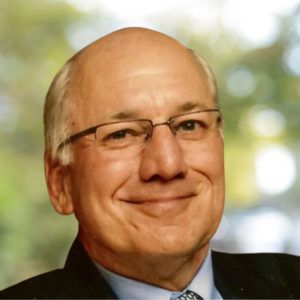 Close-up of a smiling middle-aged man with glasses, in front of a blurred outdoor background.