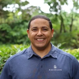 Smiling man outdoors with green trees and bushes in the background, wearing a blue collared shirt with a Mission Hope logo.