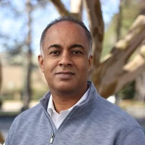 A man with short dark hair and medium skin tone, wearing a light blue zip-up jacket over a white shirt, standing outdoors with trees in the background.