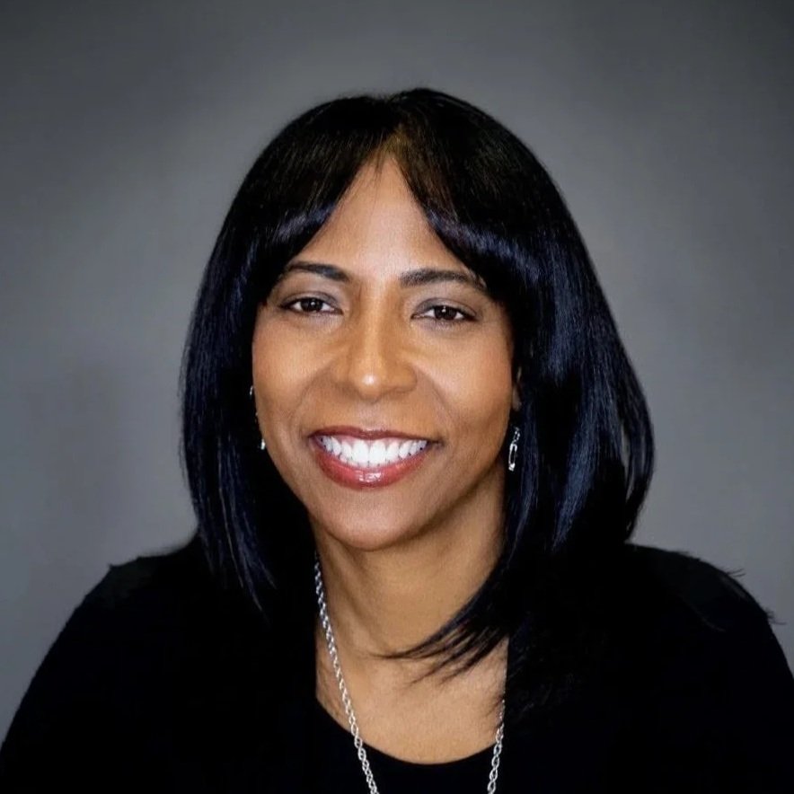 A smiling African American woman with straight black hair, wearing a black top, silver earrings, and a silver necklace, posing against a gray background.