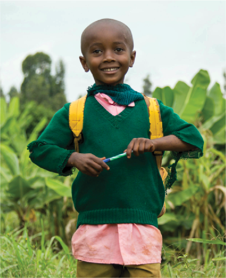 A smiling young girl in a green sweater and pink shirt outdoors in a field of banana plants, holding a pen with a yellow backpack on her shoulders.