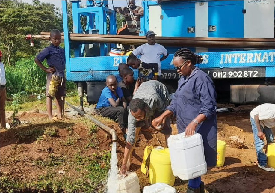A group of people collecting clean water from a pipe connected to a blue water truck, with children and women present, in an outdoor setting.