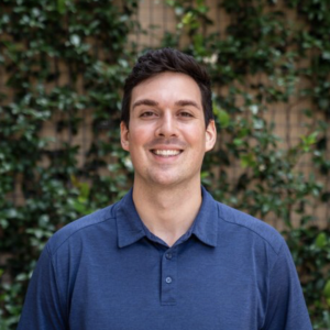 A young man with short dark hair smiling, wearing a blue polo shirt, standing outdoors with greenery and a wooden fence in the background.