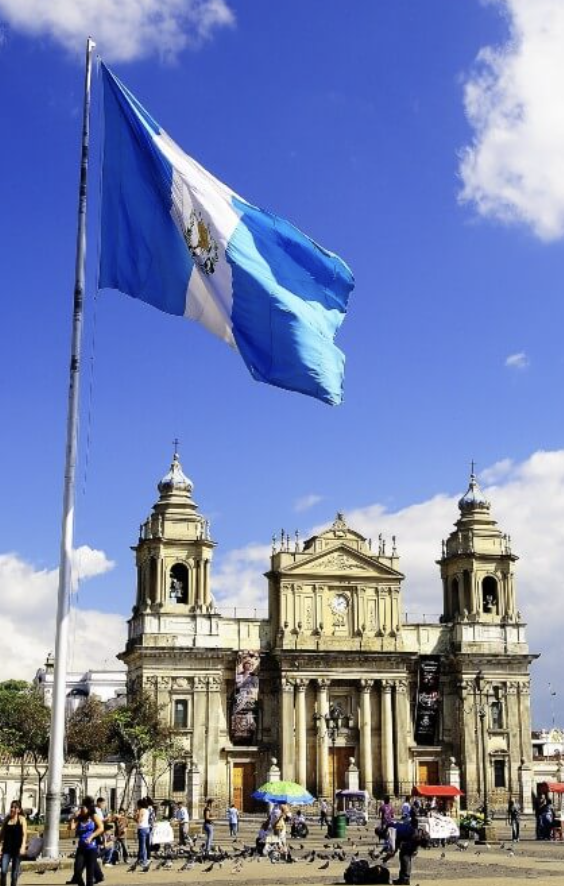Guatemalan flag flying in front of a historic church with people walking and pigeons in the plaza.